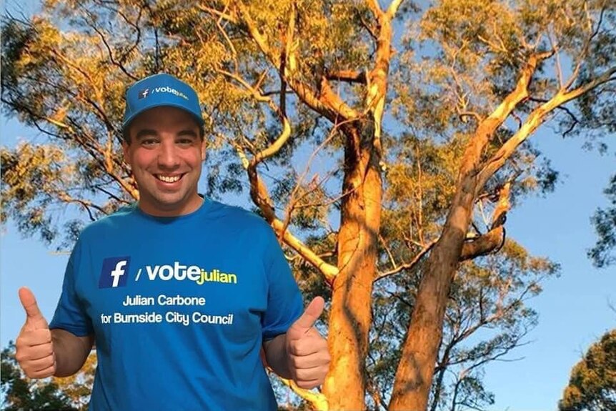 A man wearing a blue cap and campaign t-shirt with his thumbs up stands in front of trees