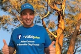 A man wearing a blue cap and campaign t-shirt with his thumbs up stands in front of trees