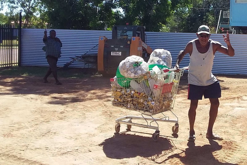 A man waves at the camera, dragging a trolley full of cans.