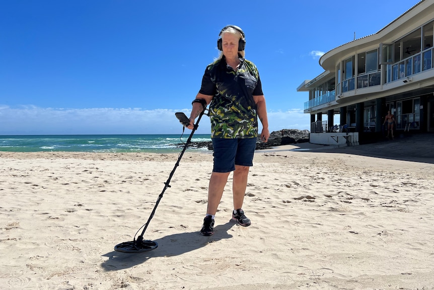 A lady with blonde hair uses a metal detector on the sand of a Gold Coast beach