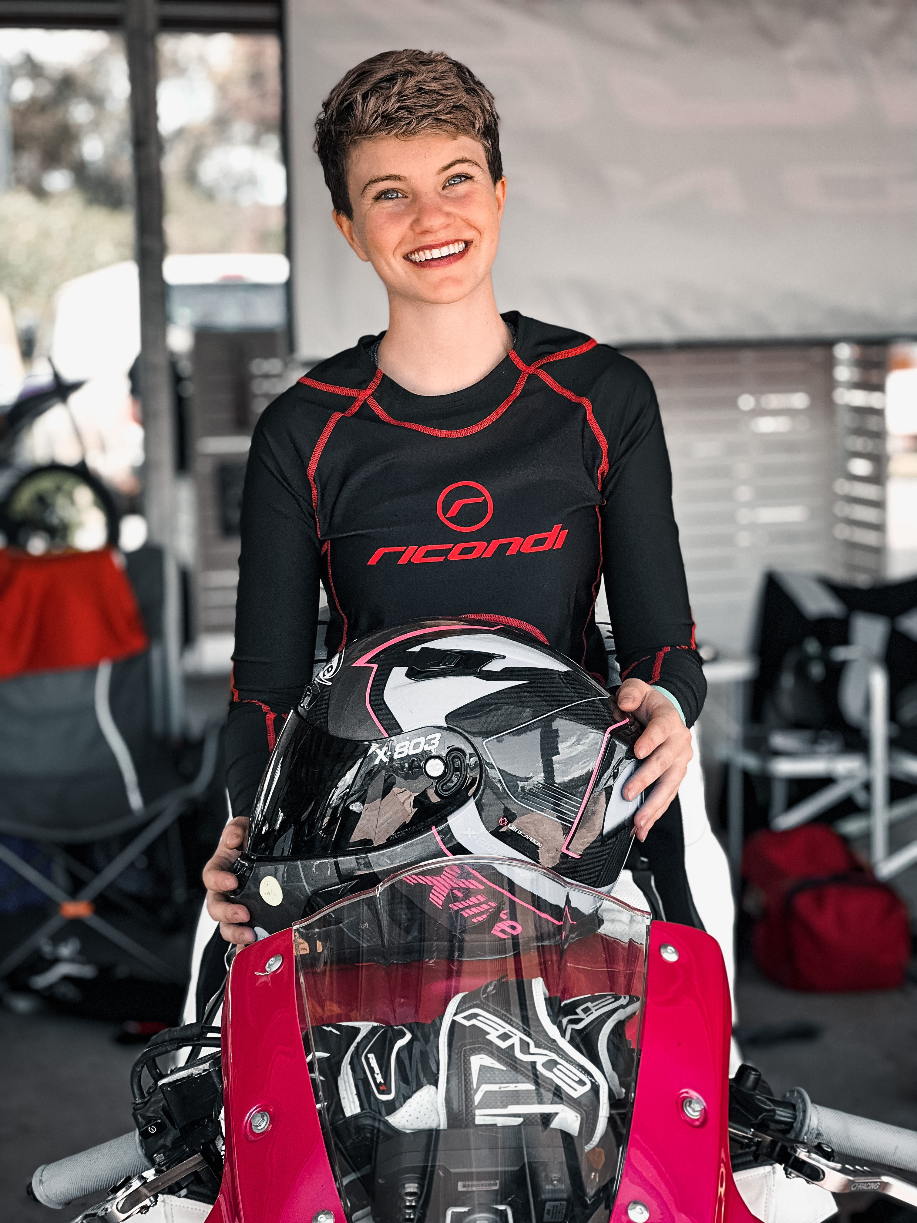 a young woman with short hair sits on a motorbike smiling, holding a black helmet