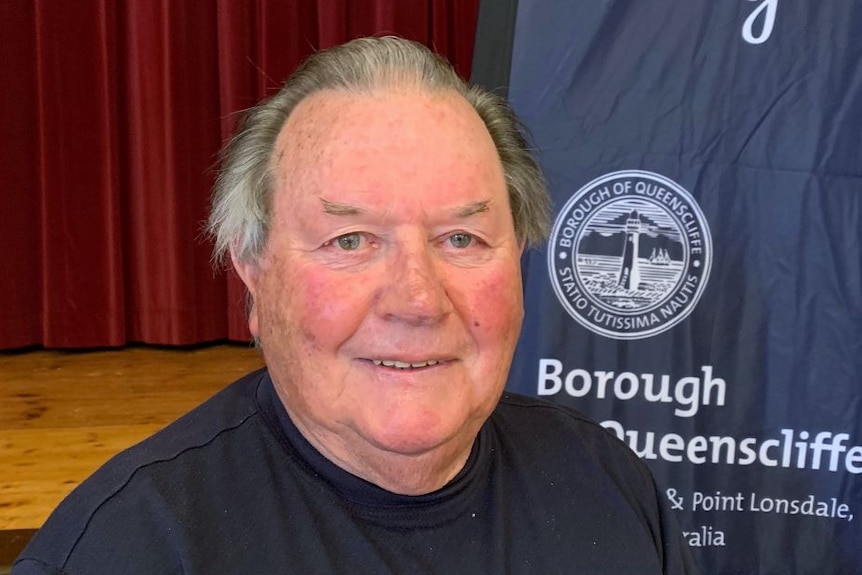 A headshot of a man standing in front of a red curtain and blue Borough of Queenscliffe flag.