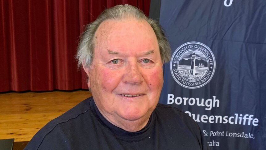 A headshot of a man standing in front of a red curtain and blue Borough of Queenscliffe flag.