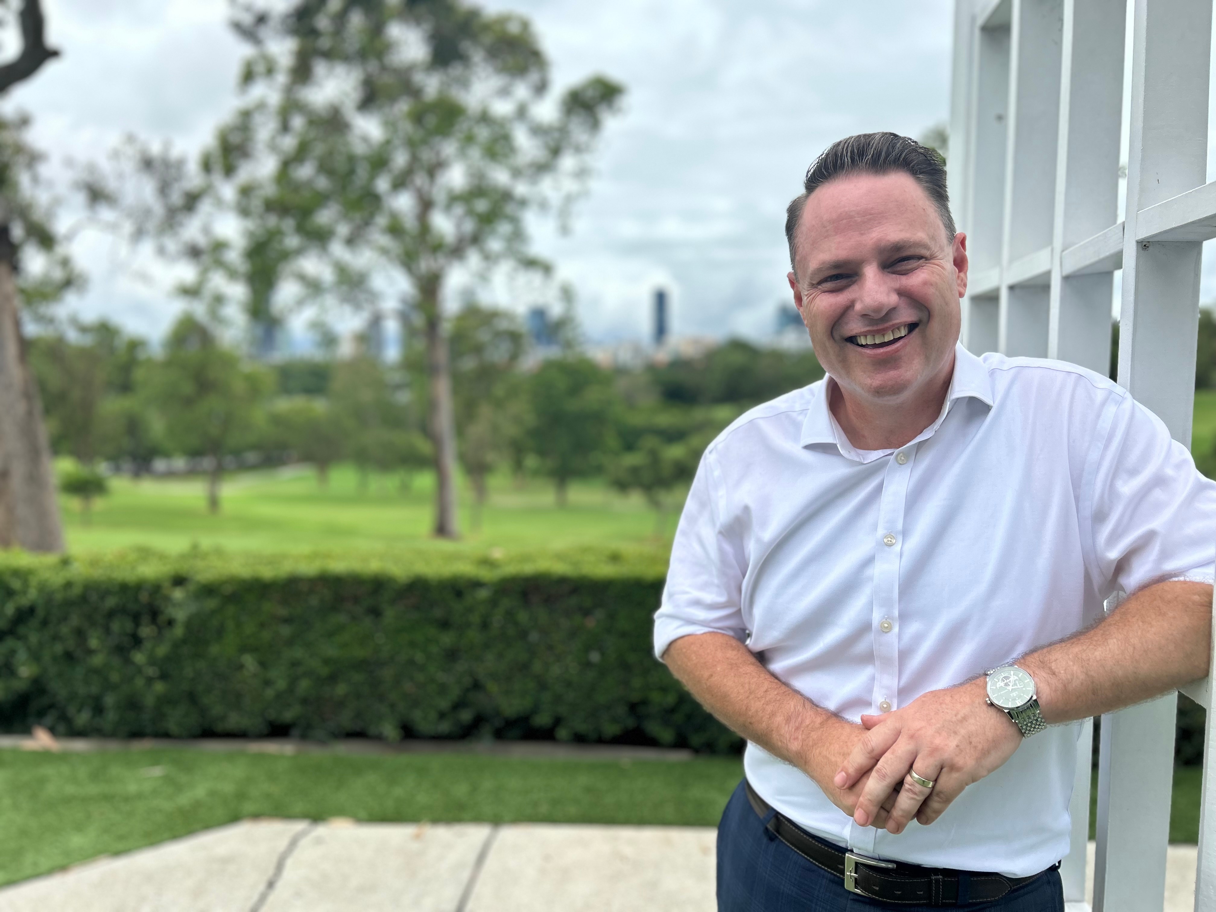 A man wearing a white business shirt leaning against a fence in front of green grass and trees.