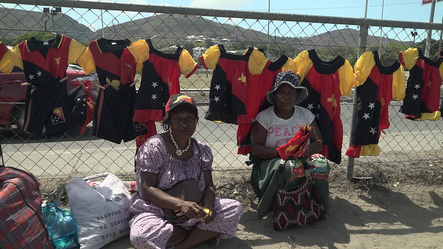 Two women sit in front of meri blouses hanging from a fence in Port Moresby.
