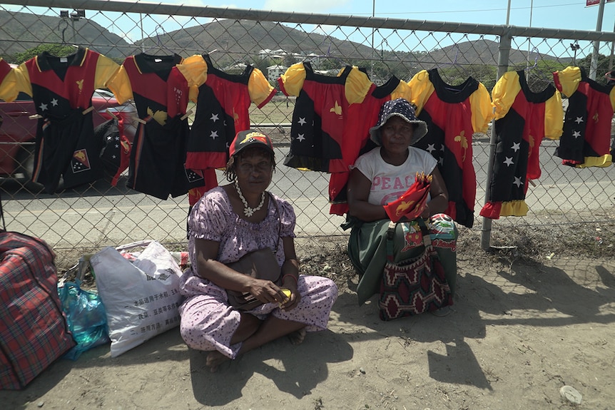 Two women sit in front of meri blouses hanging from a fence in Port Moresby.