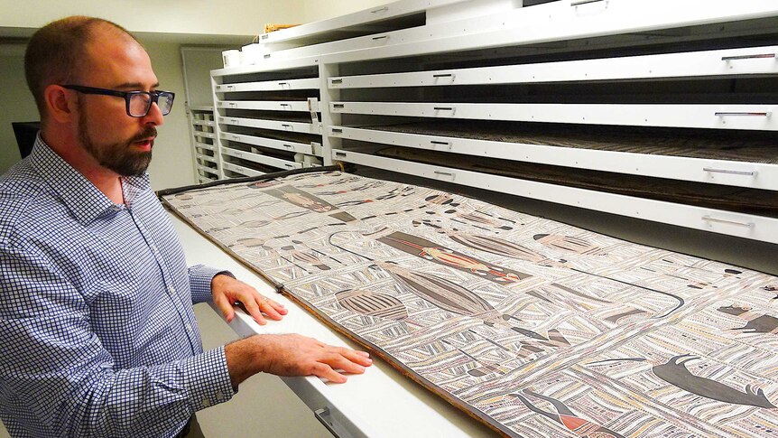 A man stands in front of a roll out drawer containing an Aboriginal painting