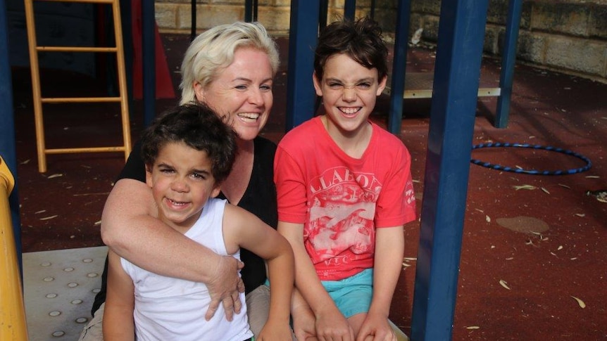 Sharon, smiling, holds her two sons on play equipment, also smiling.