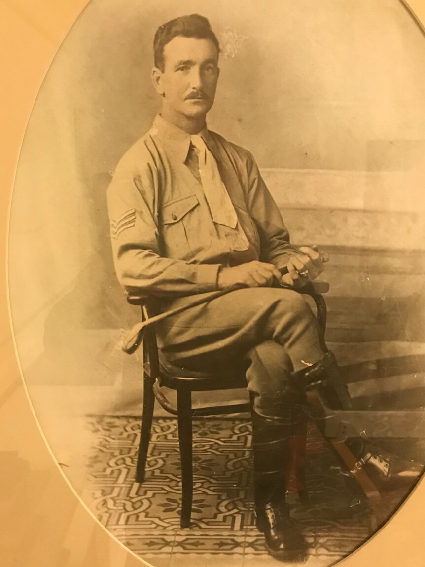 A sepia photo of a man with short hair and a mustache sitting cross legged on a wooden chair in his soldier uniform 