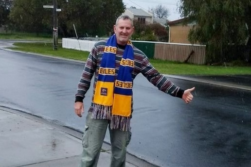 A man standing on a road holding his hand out wearing a west coast eagles scarf
