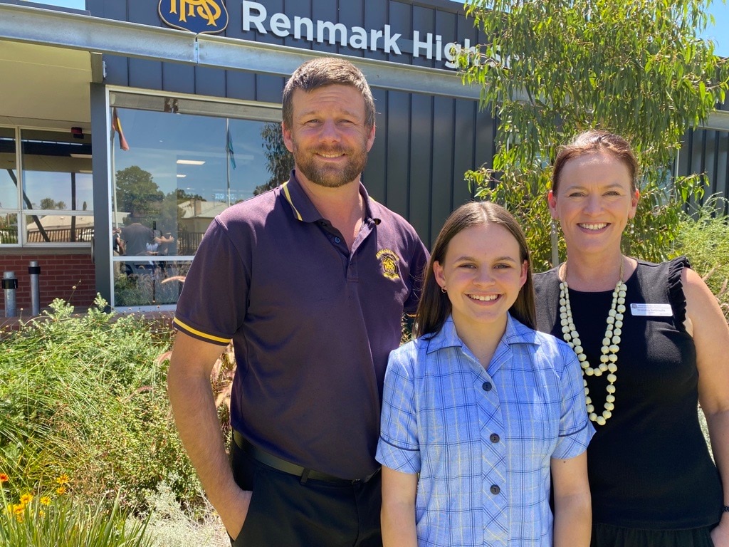 A man wearing a school teachers top smiles next to girl in blue school dress and female teacher in black top. green trees behind