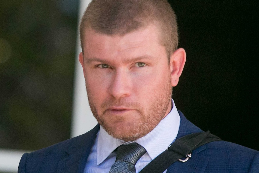 A man in a blue suit with a blue tie looks off the right side of the screen.