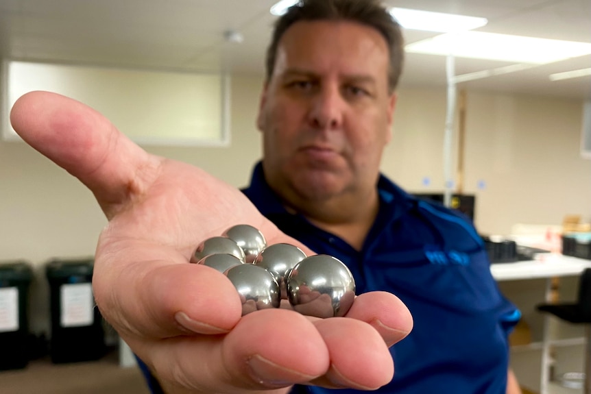 A man in a blue shirt holds out a handful of silver ball bearings close to the camera