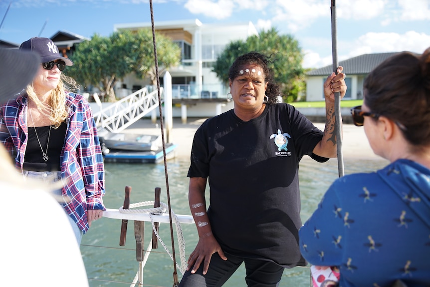 A woman in a black t-shirt with traditional white paint on her body talks to visitors aboard a ship.