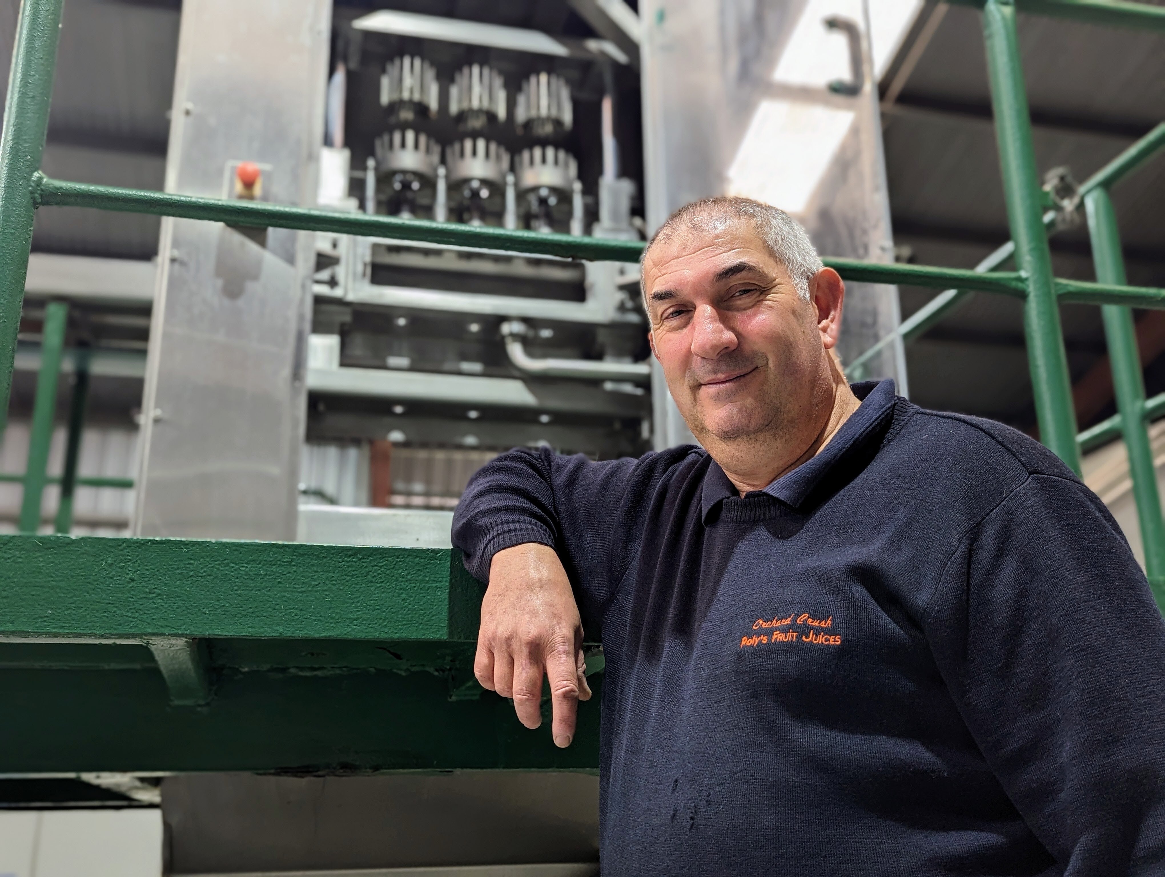 A grey-haired Greek-Australian man, smiles in front of a juice press
