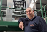 A grey-haired Greek-Australian man, smiles in front of a juice press