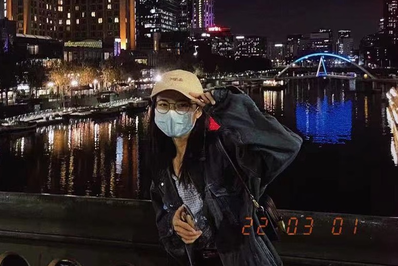 A photo of a woman in a face mask and cap at night on a bridge in lit-up city.