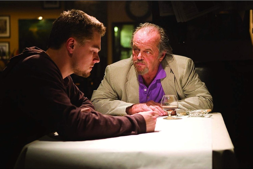 An older man looks intently at the face of a younger man, who is staring down at his hand that is stretched across a table.