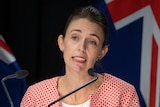 Jacinda Ardern stands at a microphone with New Zealand flags behind her