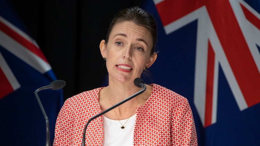 Jacinda Ardern stands at a microphone with New Zealand flags behind her