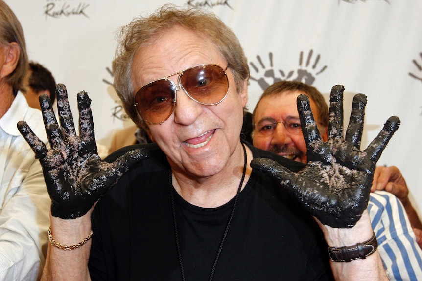 Hal Blaine smiles in midshot as he holds his hands up with cement on them. He is wearing sunglasses, and a black t shirt.