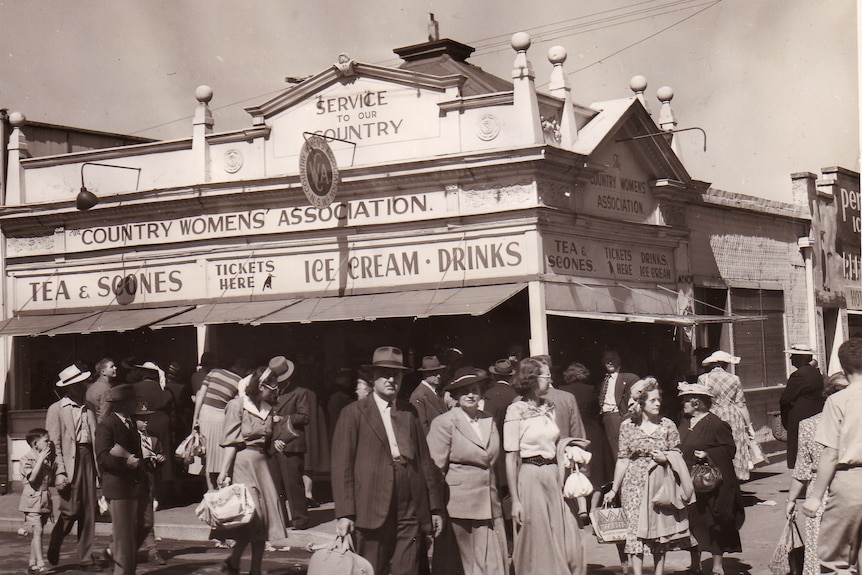 Men, women and children pass a building corner with Country Women's Association painted on.