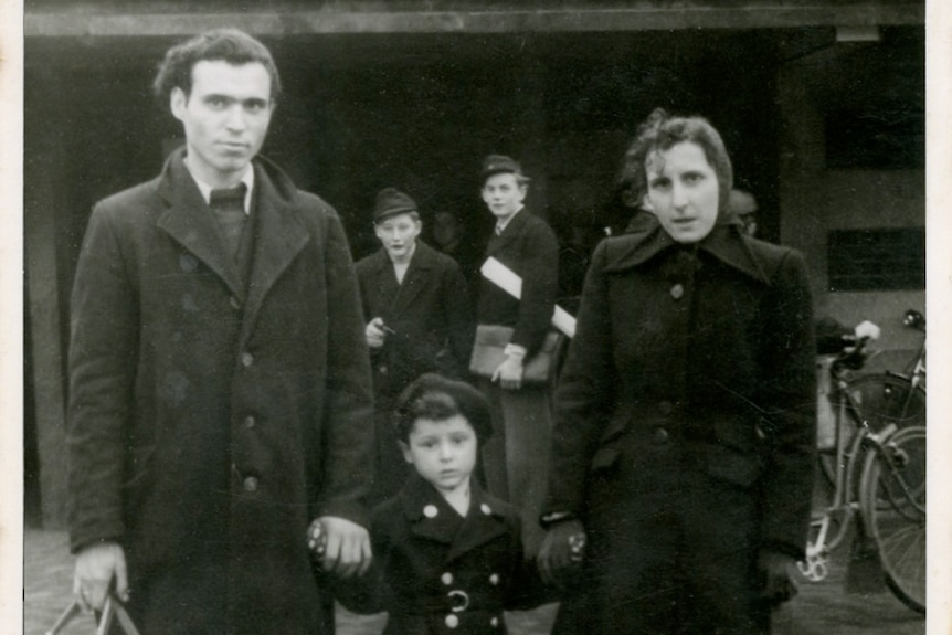A black and white photo of very young Rai holding hands with his mother and father. Two other figures also look at the camera.