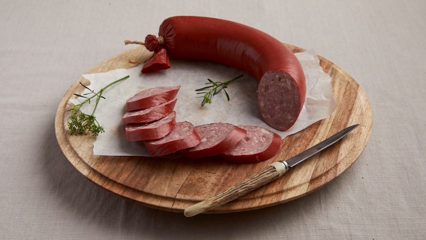 A red sausage on a board partly sliced and partly whole with a knife and parsley
