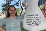 A woman stands next to a cardboard dress that says 'Over 600,000 Australian women are at risk of homelessness' 