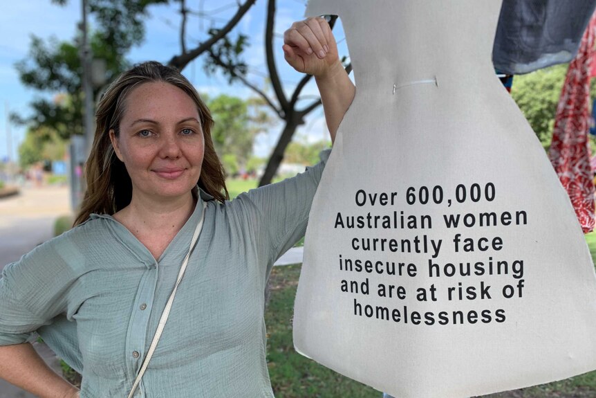 A woman stands next to a cardboard dress that says 'Over 600,000 Australian women are at risk of homelessness' 