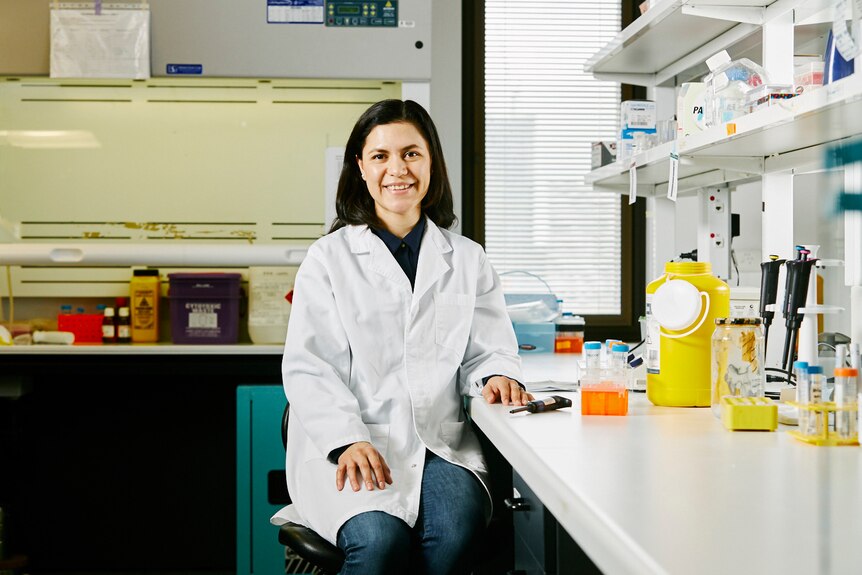 Smiling woman with dark shoulder length hair wearing a white lab coat sitting in a laboratory  