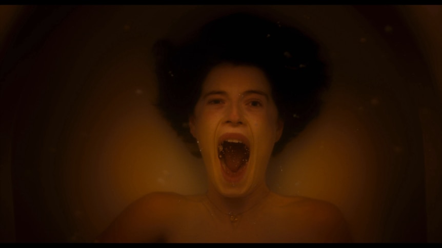 White woman with short dark hair screams from the inside of a bathtub with her head underwater and eyes open in terror.