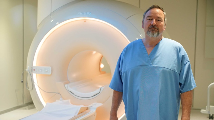 A middle-aged man in a hospital gown stands next to a CT machine
