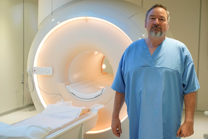 A middle-aged man in a hospital gown stands next to a CT machine