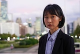 A close up of a woman with black hair wearing a suit in frotn of a city.