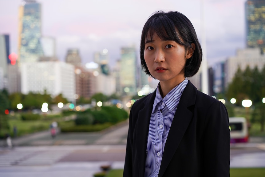 A close up of a woman with black hair wearing a suit in frotn of a city.