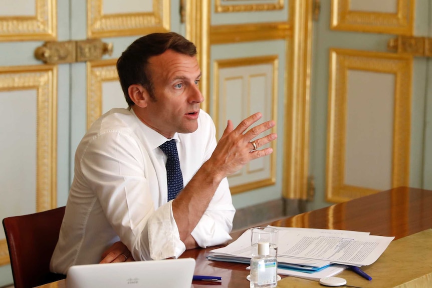 Macron has rolled up white sleeves and gestures as he talks while sitting at a desk in a room with gold trimmings.
