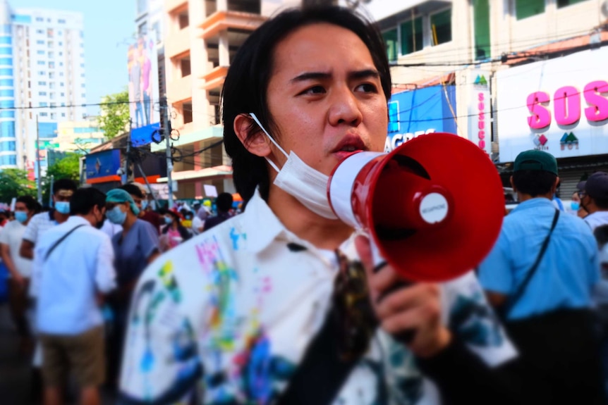 A man with a mask on holds a megaphone to his mouth as he looks out over a crowd of protesters on a street.