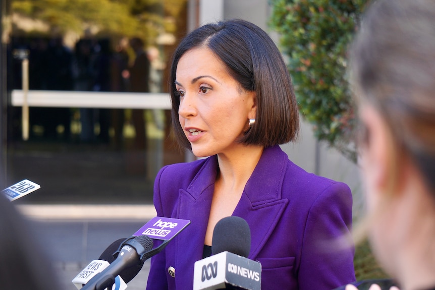 A woman with dark hair, wearing a purple jacket, speaking into microphones.