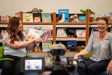 Two women in arms chairs read a children's book in front of a video camera