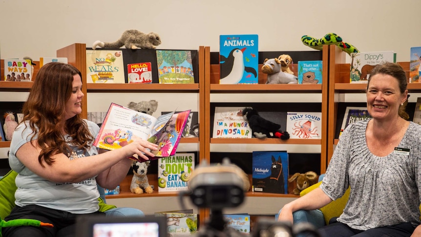 Two women in arms chairs read a children's book in front of a video camera