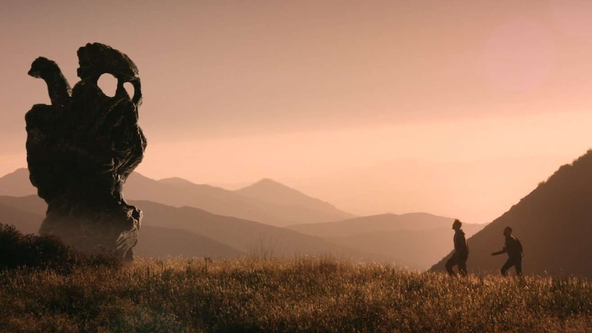 Still image from 2017 The Endless, two characters in a dusty, hilly and mountainous area approach a large rock like object.