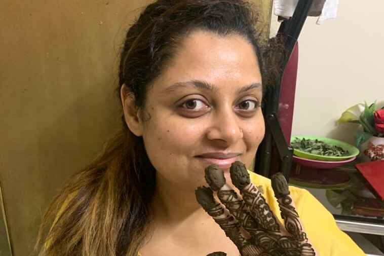 A women smiles at the camera, holding up her hand which has a raw henna design on it.