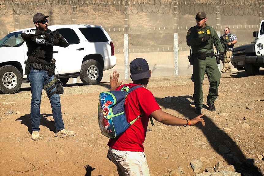 A man holds up his hand as two agents with guns and protective gear direct him to turn around as they stand in front of fence.