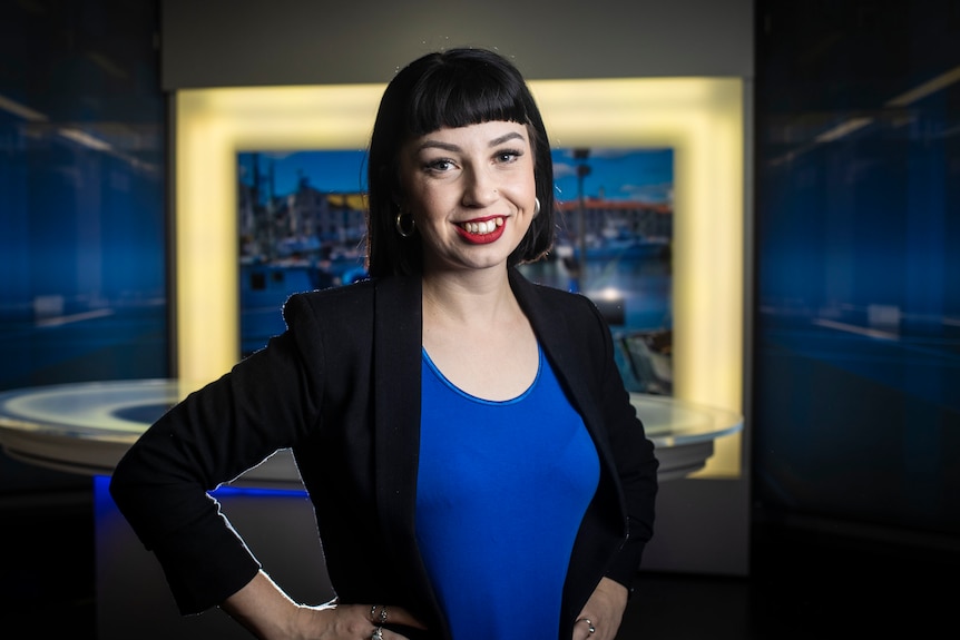 Emily has black hair and is dressed in blue with a black blazer and stands in a television news studio