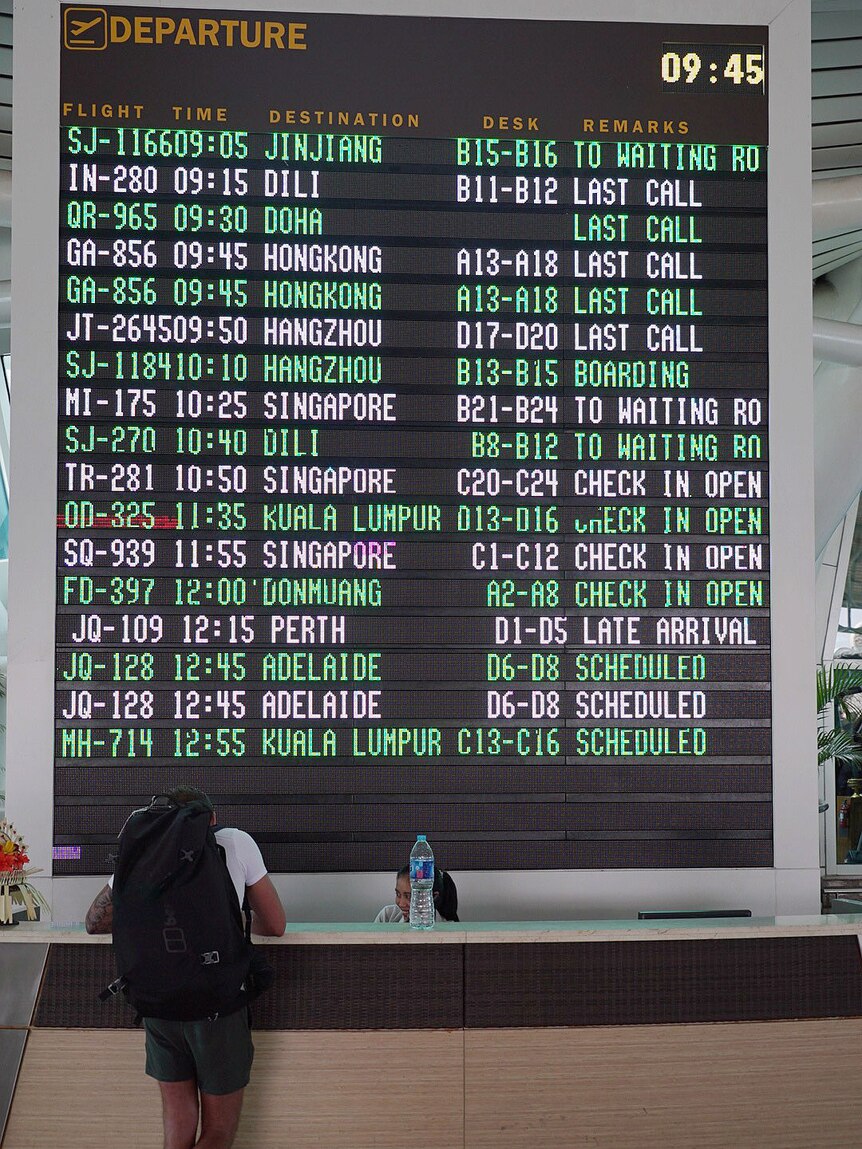 A man leans on an airport desk in front of a departure board.