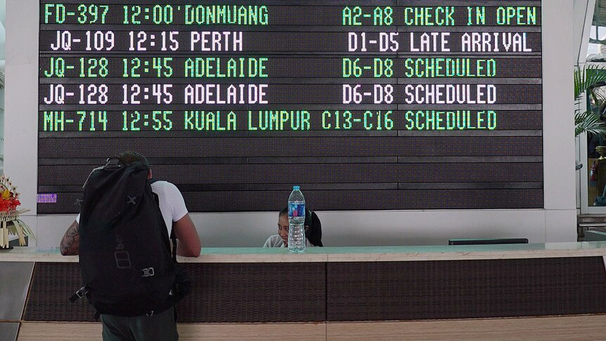 A man leans on an airport desk in front of a departure board.
