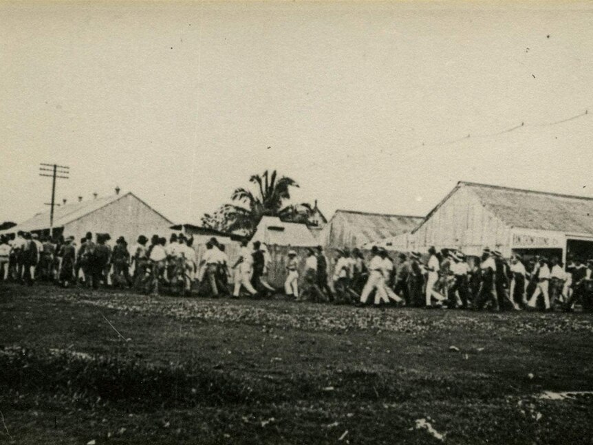 Black and white image of a line of men marching around a corner with small old buildings in the background.