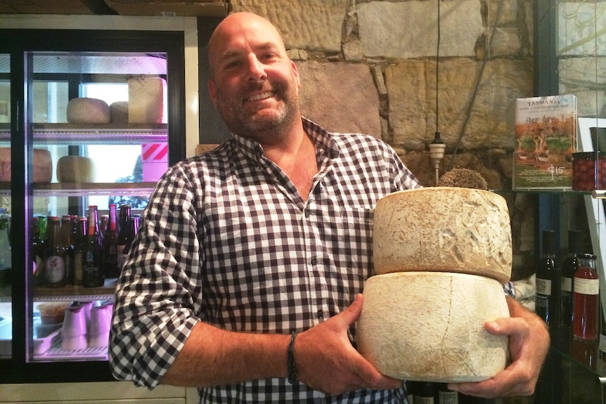 A man smiles while holding two big wheels of cheese.