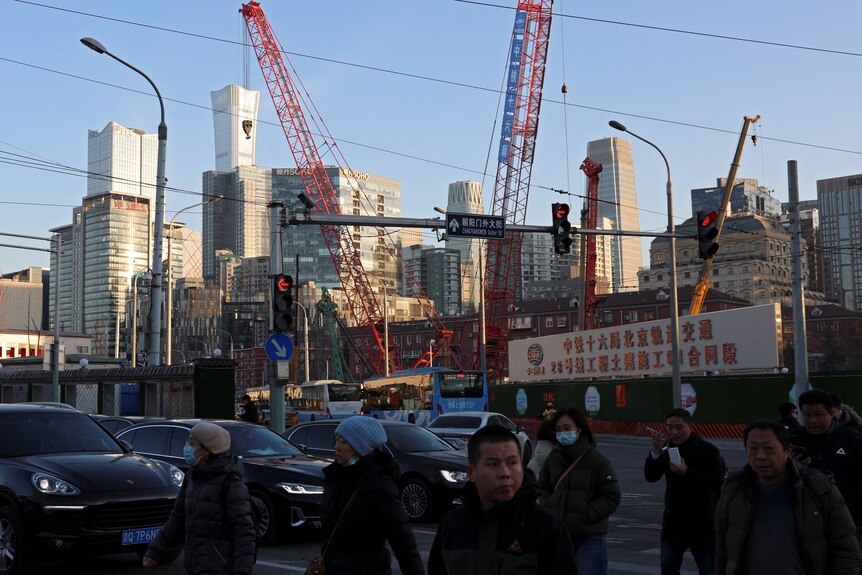 People cross at an intersection with traffic lights and cars. In the distance are high rise buildings and cranes.